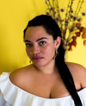 Ama Josephine Budge, a brown-skinned person with long black braided hair stands in front of a yellow background looking into the camera. Framed from the chest up, Budge is wearing a white blouse, a round nose ring, and earrings.