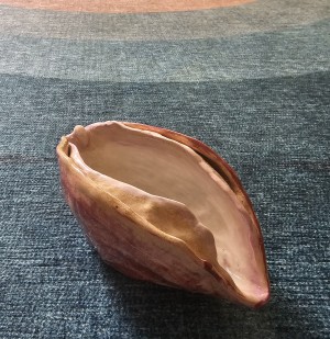 A ceramic bowl shaped like a vulva, colored with different hues of pink and red is placed on a blue textile surface. 