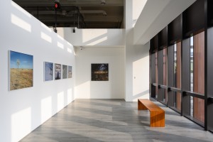 Exhibition view of a light space with big windows on the right side. There are six photographs on the white wall, some depicting landscapes with hay and blue sky, some photographs further back have people and built environment depicted in them. 