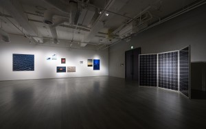 Exhibition room with an object that looks like a solar panel, and photographs further on the back wall. 