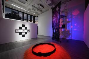 Exhibition view. In the front there is a bright red dome of sand-like material. On the left three photographs and six black rectangulars of the size of those photographs form a diamond shape. Next to the back wall is a tall, sparkly, metallic curtain attached to a high scaffolding on wheels, with a pink and blue shadow of the structure reflected on the wall. 