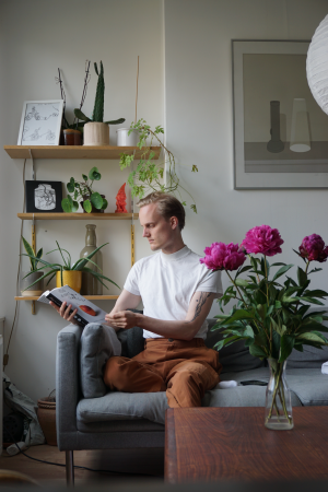 Oskar Koli, valkoihoinen, lyhythiuksinen ja vaaleatukkainen henkilö, istuu sohvalla lukien kirjaa. Kolilla on yllään valkoinen paita ja oranssit housut.